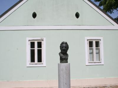 A light green single-story building with white trim and two windows, featuring a bronze bust of a man on a stone pedestal in the foreground.