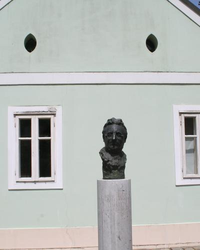 A light green single-story building with white trim and two windows, featuring a bronze bust of a man on a stone pedestal in the foreground.