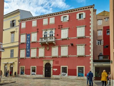 A tall, salmon-pink baroque building with white shutters and a prominent vertical blue banner reading "MUSEUM" hanging on its facade, situated in a paved European square.