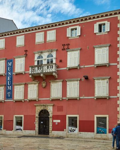 A tall, salmon-pink baroque building with white shutters and a prominent vertical blue banner reading "MUSEUM" hanging on its facade, situated in a paved European square.