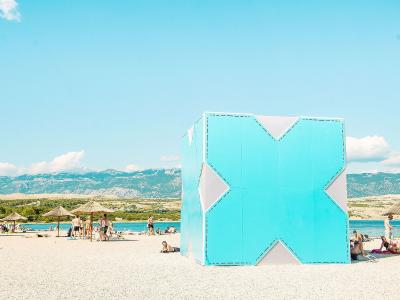 A large, light-blue geometric cube structure with white diamond cutouts stands on a white pebble beach, with people relaxing under straw umbrellas and mountains in the background.