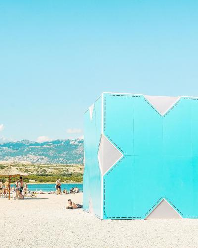 A large, light-blue geometric cube structure with white diamond cutouts stands on a white pebble beach, with people relaxing under straw umbrellas and mountains in the background.