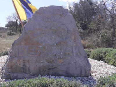 A large, rough-hewn stone monument engraved with the word "DEBELJAK" sits on a bed of white gravel, with a yellow and blue flag flying to the left and trees in the background.