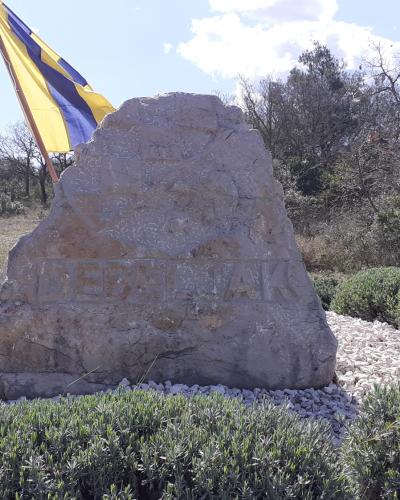 A large, rough-hewn stone monument engraved with the word "DEBELJAK" sits on a bed of white gravel, with a yellow and blue flag flying to the left and trees in the background.
