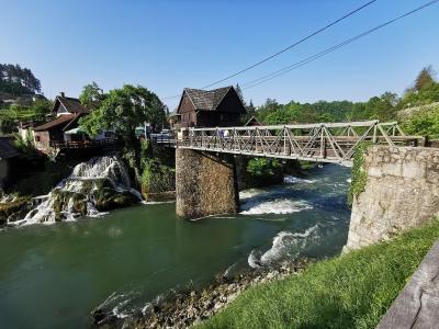A metal truss bridge spanning a river with cascading waterfalls and traditional wooden buildings on the banks under a clear blue sky.