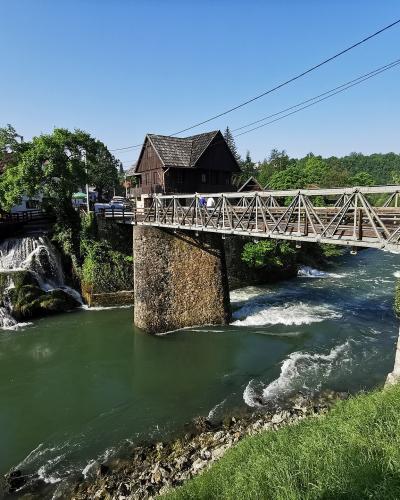 A metal truss bridge spanning a river with cascading waterfalls and traditional wooden buildings on the banks under a clear blue sky.