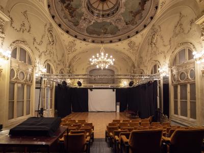 An ornate interior hall with a painted dome ceiling, chandeliers, and arched windows, set up with rows of chairs facing a temporary stage and projection screen.