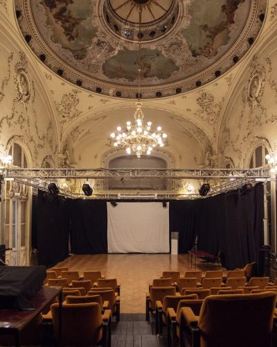 An ornate interior hall with a painted dome ceiling, chandeliers, and arched windows, set up with rows of chairs facing a temporary stage and projection screen.