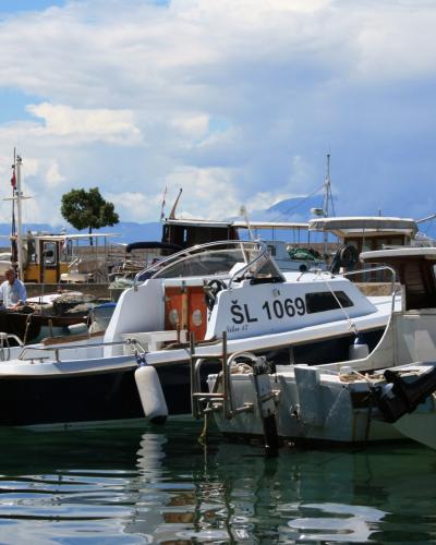 A daytime view of several small motorboats and fishing vessels moored in a harbor, with a person visible on a dock in the background.