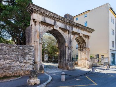 A photograph of the Porta Gemina, a double-arched Roman triumphal gate in Pula, Croatia, featuring weathered stone columns and an entablature with decorative molding, set against a modern building and trees.