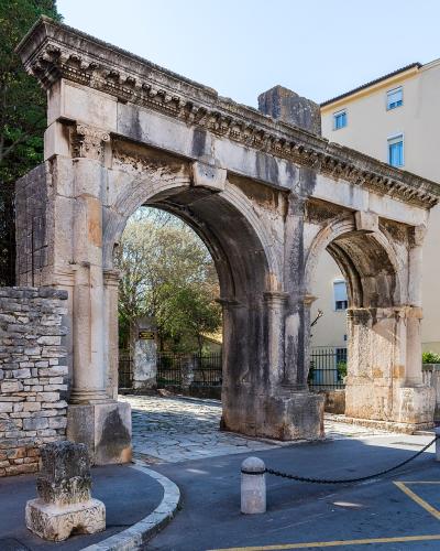 A photograph of the Porta Gemina, a double-arched Roman triumphal gate in Pula, Croatia, featuring weathered stone columns and an entablature with decorative molding, set against a modern building and trees.
