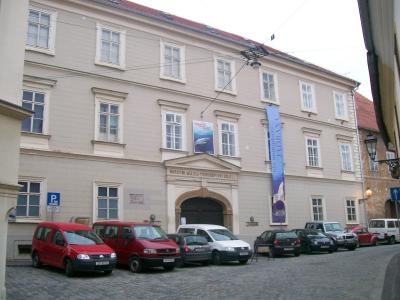 A beige, multi-story historic building with a central arched entrance, displaying a sign above the door that reads "NARODNI MUZEJ PRIRODOPIŠNI ODJELI" and a vertical blue banner on the right side; several cars are parked on the cobblestone street