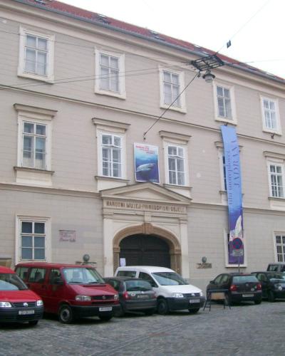 A beige, multi-story historic building with a central arched entrance, displaying a sign above the door that reads "NARODNI MUZEJ PRIRODOPIŠNI ODJELI" and a vertical blue banner on the right side; several cars are parked on the cobblestone street
