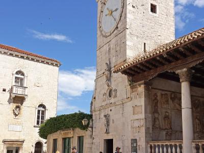 A low-angle shot of a tall, white stone clock tower with a red-tiled pyramidal roof and a large clock face, situated in a paved public square with outdoor cafe seating and adjacent historic buildings under a blue sky.