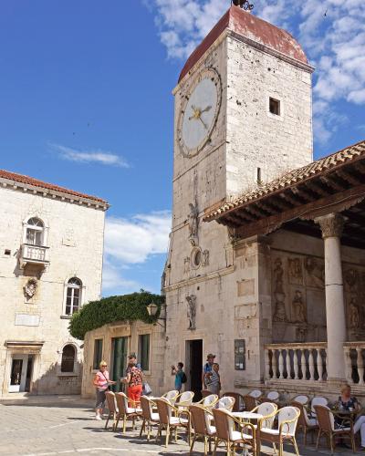 A low-angle shot of a tall, white stone clock tower with a red-tiled pyramidal roof and a large clock face, situated in a paved public square with outdoor cafe seating and adjacent historic buildings under a blue sky.