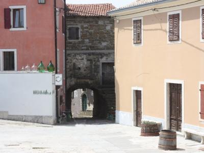 A sunlit stone-paved square in a European hilltop town, featuring a pink building on the left, a beige building on the right, and a stone archway leading to a narrow passage in the center background.