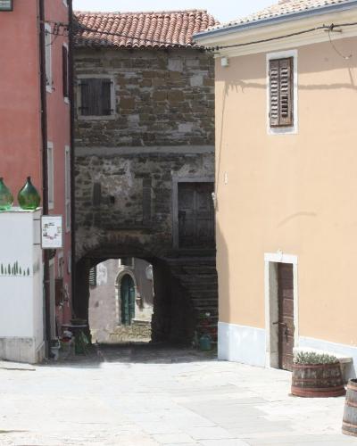 A sunlit stone-paved square in a European hilltop town, featuring a pink building on the left, a beige building on the right, and a stone archway leading to a narrow passage in the center background.