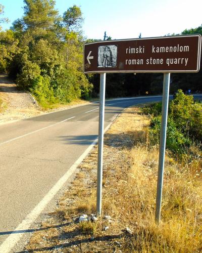 A brown tourist sign on a roadside in a wooded area reads "rimski kamenolom" and "roman stone quarry" with an arrow pointing left.