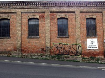 A red brick building facade with four arched windows, graffiti on the lower wall, and a white sign reading "GLIPTOTEKA" with a logo above it.
