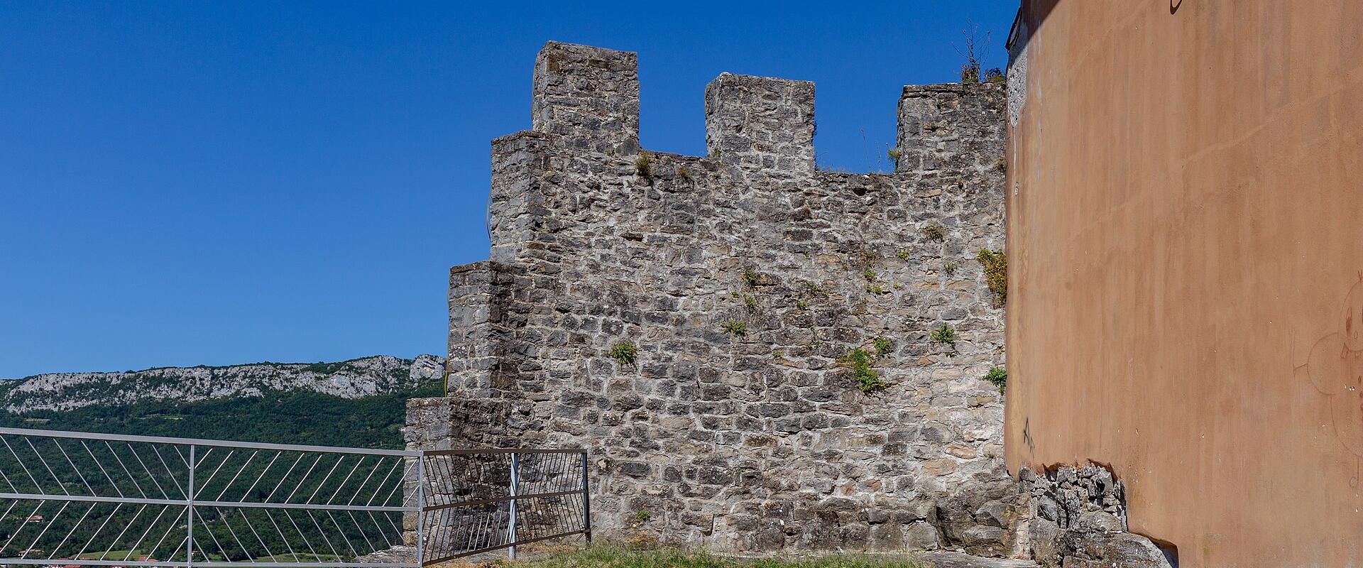 A section of ancient stone fortification walls with crenellations, adjacent to a terracotta-colored building and a modern metal railing, overlooking a green valley and distant hills under a clear blue sky.