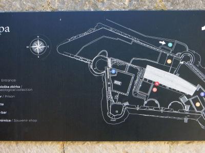 A close-up photograph of a black informational map sign mounted on a stone wall, displaying a site plan with numbered points of interest and a logo in the top right corner.