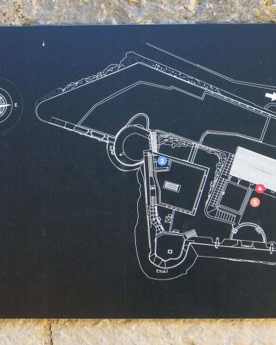 A close-up photograph of a black informational map sign mounted on a stone wall, displaying a site plan with numbered points of interest and a logo in the top right corner.