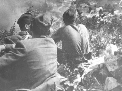A black and white historical photograph showing three soldiers in military uniforms and caps from behind, overlooking a village in a valley with smoke rising in the distance.