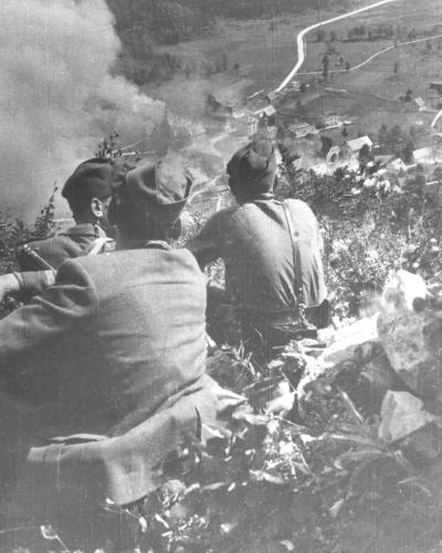 A black and white historical photograph showing three soldiers in military uniforms and caps from behind, overlooking a village in a valley with smoke rising in the distance.