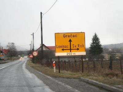 A yellow directional road sign on a wet roadside indicating directions to Gračac (straight), Lovinac (left), and Sv. Rok (right) in the Lika region of Croatia.