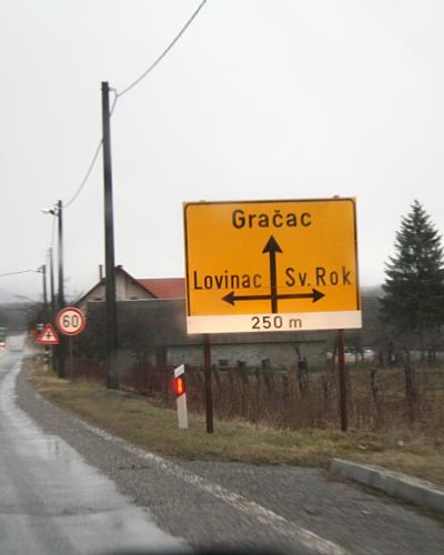 A yellow directional road sign on a wet roadside indicating directions to Gračac (straight), Lovinac (left), and Sv. Rok (right) in the Lika region of Croatia.