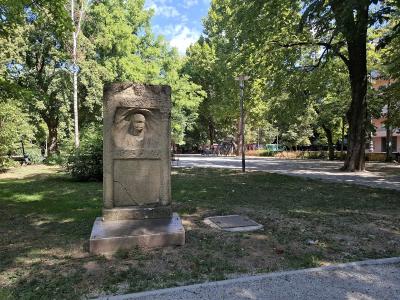 A stone monument featuring a relief portrait of a man stands in a grassy, tree-filled park with a paved path and playground equipment visible in the background under a blue sky.