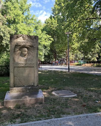 A stone monument featuring a relief portrait of a man stands in a grassy, tree-filled park with a paved path and playground equipment visible in the background under a blue sky.