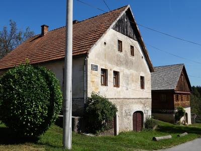 A two-story rural house with a tiled roof, white stucco facade, and a small plaque on the wall, situated next to a wooden outbuilding under a clear blue sky.