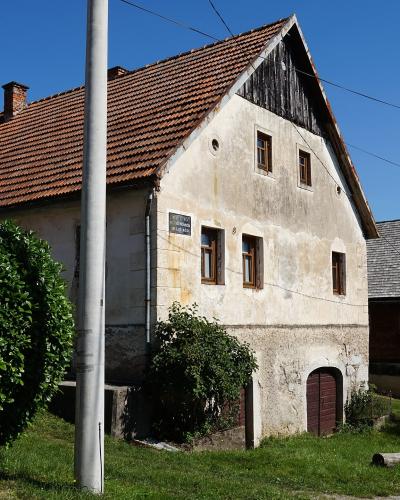 A two-story rural house with a tiled roof, white stucco facade, and a small plaque on the wall, situated next to a wooden outbuilding under a clear blue sky.