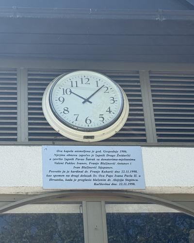 A close-up of a building facade showing a round clock and a commemorative plaque with Croatian text detailing the history and consecration of a chapel in Kučilovina.