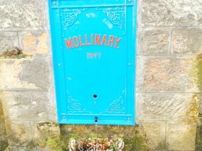 A close-up of a bright blue, ornate metal water fountain panel mounted on a stone wall, featuring the red text "MOLLINARY." and the date "1877" above a spout and stone basin.