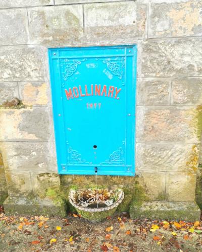 A close-up of a bright blue, ornate metal water fountain panel mounted on a stone wall, featuring the red text "MOLLINARY." and the date "1877" above a spout and stone basin.