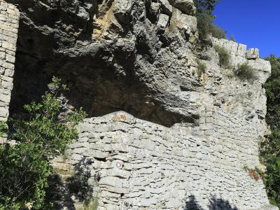 A low-angle view of a rocky cliff face with a stone retaining wall and steps leading up to a cave entrance, set against a clear blue sky.