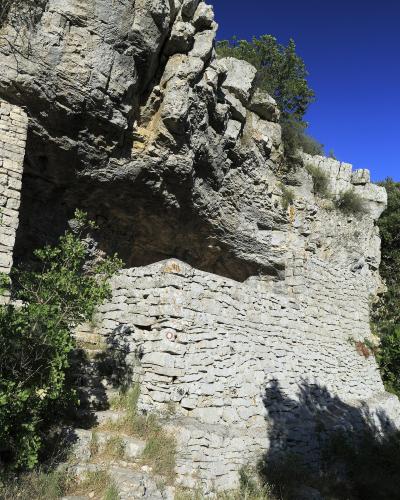A low-angle view of a rocky cliff face with a stone retaining wall and steps leading up to a cave entrance, set against a clear blue sky.