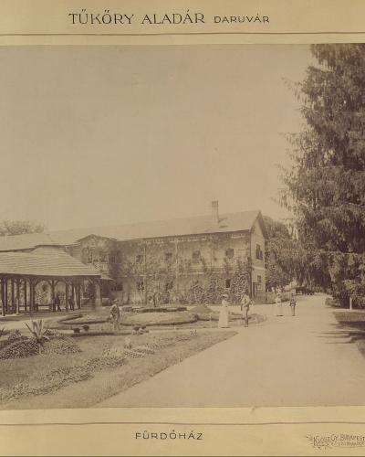 A sepia-toned historical photograph showing a large, ivy-covered building with a veranda and garden, identified by text at the top as "TÜKÖRY ALADÁR DARUVÁR" and at the bottom as "FÜRDŐHÁZ" (Bath House).
