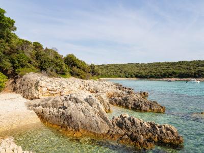A rocky, pebbled cove with clear turquoise water, bordered by dense green Mediterranean vegetation and a small sandy beach area, with boats visible in the distance.