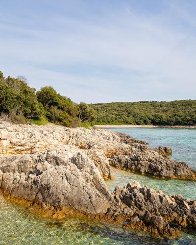 A rocky, pebbled cove with clear turquoise water, bordered by dense green Mediterranean vegetation and a small sandy beach area, with boats visible in the distance.