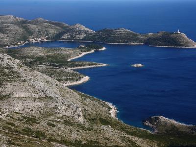 A high-angle landscape view of a rugged, rocky coastline featuring a sheltered bay with a small settlement of buildings with red roofs, and a prominent lighthouse on a headland to the right.