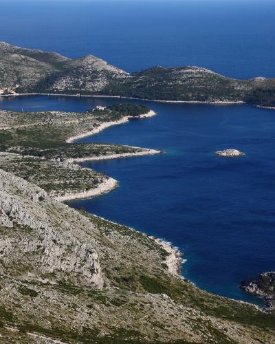 A high-angle landscape view of a rugged, rocky coastline featuring a sheltered bay with a small settlement of buildings with red roofs, and a prominent lighthouse on a headland to the right.