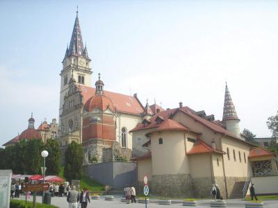 The image shows a large, ornate church complex with a prominent tall spire featuring a patterned roof, red-tiled roofs on adjacent buildings, and a plaza with people, vendor stalls, and signage in the foreground.