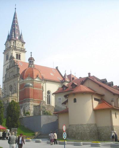 The image shows a large, ornate church complex with a prominent tall spire featuring a patterned roof, red-tiled roofs on adjacent buildings, and a plaza with people, vendor stalls, and signage in the foreground.