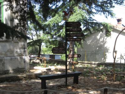 A stone-paved outdoor area with a wooden bench, a signpost featuring a cartoon bird and directional signs pointing to "Beli", "Ornitološki rezervat 'Kruna'", and "Grad Cres", and a stone building in the background.