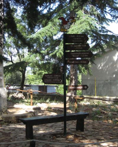 A stone-paved outdoor area with a wooden bench, a signpost featuring a cartoon bird and directional signs pointing to "Beli", "Ornitološki rezervat 'Kruna'", and "Grad Cres", and a stone building in the background.