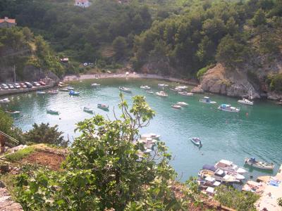 A high-angle view of a sheltered cove filled with numerous small boats and yachts, with a small pebble beach and parking area visible on the left shore, surrounded by green, hilly terrain.