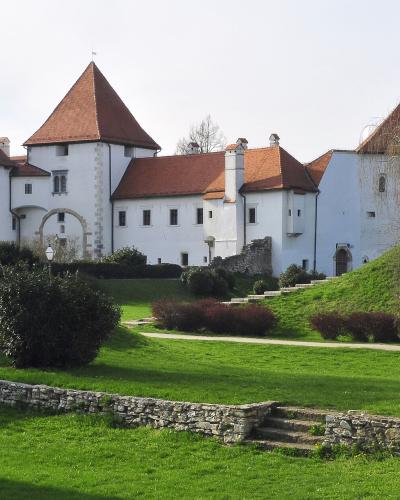 A wide shot of a large, white-walled castle complex with distinctive red-tiled conical roofs and towers, set on a grassy hill with stone ruins in the foreground.